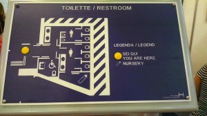 Mapa tátil de sanitário público com fundo aziul escuro e paredes em branco. Detalha localização dos sanitários feminino, masculino, acessível e elementos internos