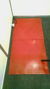 Foto mostra duas plascas de piso tátil no chão do estande. Uma com raias em curva e a outra com relevos em bolinha pequenas e próximas. As placas tem cor vermelha.