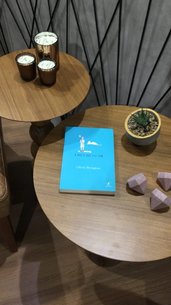 Foto vista de cima das mesas de apoio em duas alturas, com o livro "O que é que ele tem" da Olivia Byington, vela, vaso e detalhes decorativos