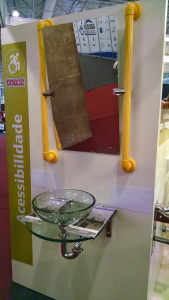 Foto de pia com espelho no estande. São duas barras verticais amarelas nas laterais, que suportam do espelho por um eixo central.